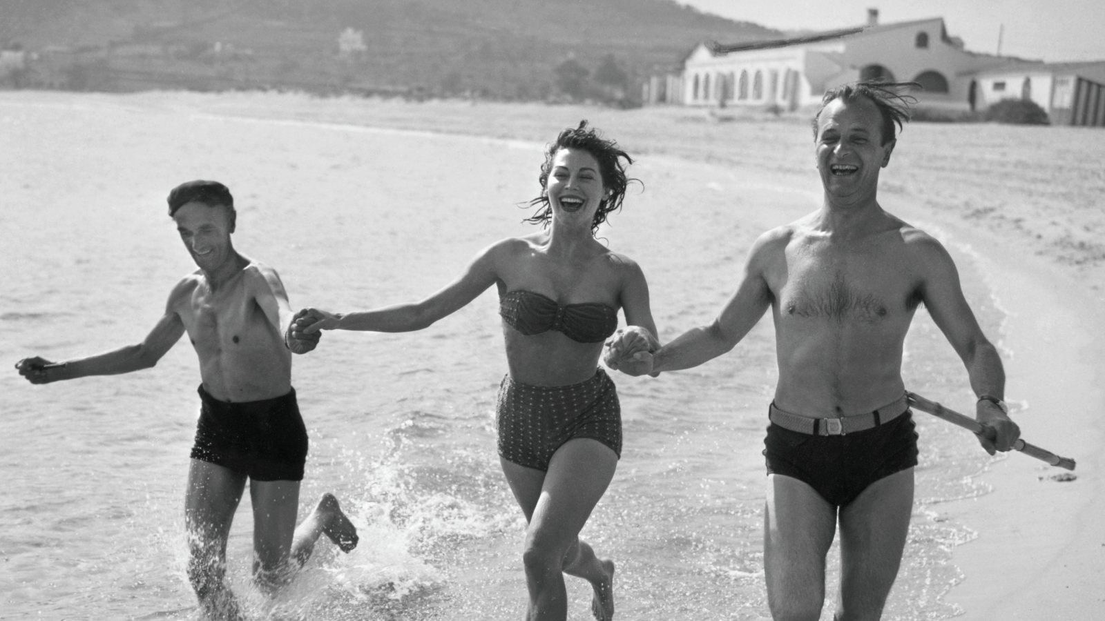 Ava Gardner con John Laurie (a su derecha) y Nigel Patrick, miembros del reparto de Pandora y el holandés errante (1951), corriendo por la playa de Sant Pol, con el restaurante La Taverna del Mar, al fondo.
